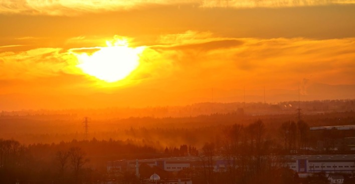 Sonnenuntergang mit glutrotem Himmel über einer bewaldeten Landschaft und Industriegebäuden, Hitze- und Dunstschleier sichtbar.