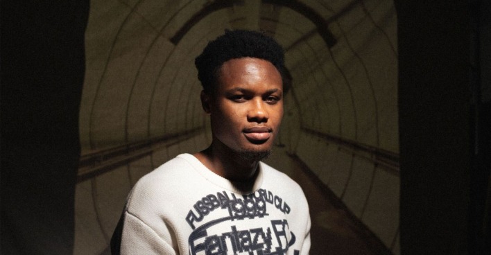 Kelvin Boateng vor einer Leinwand sitzend, welche den Durchgang der U-Bahn-Station Altes Landgut zeigt