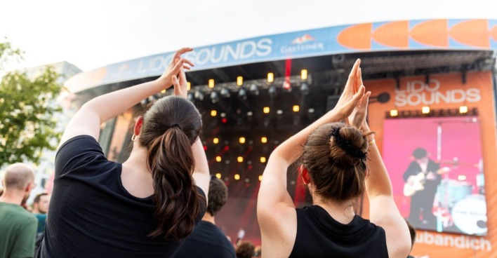 Zwei junge Frauen klatschen und feiern in einer Menschenmenge vor der Lido-Sounds-Bühne in Linz. Im Hintergrund sind bunte Lichter, ein LED-Screen mit Live-Musik und das Logo des Festivals zu sehen.
