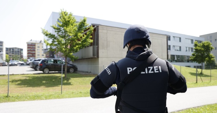 Ein Polizist in Schutzkleidung und mit Helm steht vor einem modernen Schulgebäude. Im Hintergrund sind parkende Autos und weitere Gebäude sichtbar. Die Szene spielt bei Tageslicht im Freien, das Gelände ist eingezäunt.