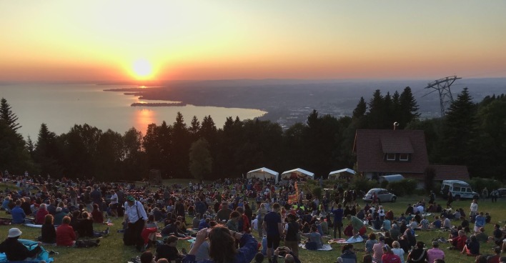 Menschen sitzen auf einer Wiese am Pfänder und blicken auf den Bodensee bei Sonnenuntergang während des Sonnwend-Fests mit Feuerstelle und Festzelten.