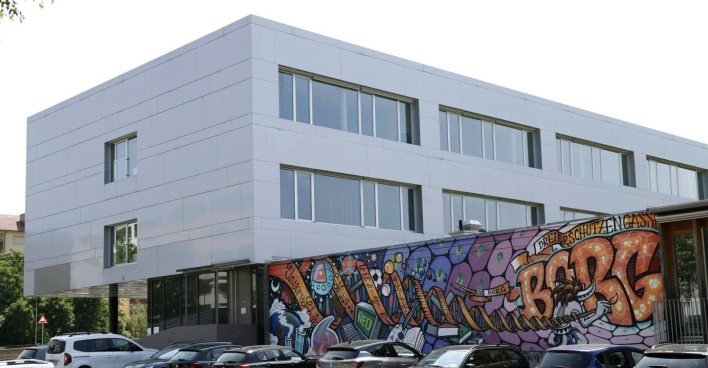Modernes Schulgebäude mit weißer Fassade und großem Graffiti mit der Aufschrift „BORG Dreierschützengasse“ an der Frontwand. Mehrere Autos sind davor geparkt.
