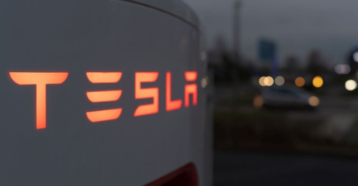 Leuchtender Tesla-Schriftzug an einer Ladesäule bei Dämmerung, im Hintergrund unscharfe Straßenlichter und Autos.. Nach dem öffentlichen Streit zwischen Elon Musk und Donald Trump stürzt die Tesla-Aktie ab.