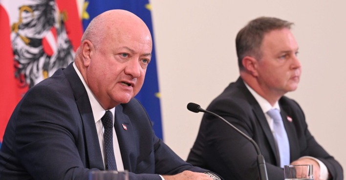 Christian Stocker und Andreas Babler sitzen bei einer Pressekonferenz vor Mikrofonen. Im Hintergrund sind die Flaggen Österreichs und der EU zu sehen.