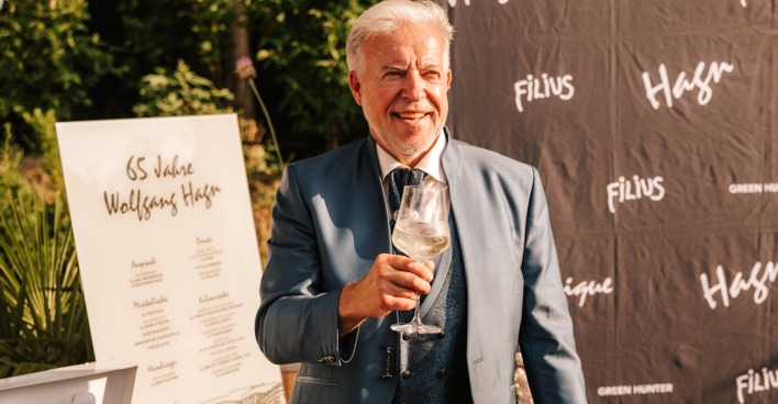 Mann mit weißen Haaren im grauen Festagsanzug und einem Glas Wein