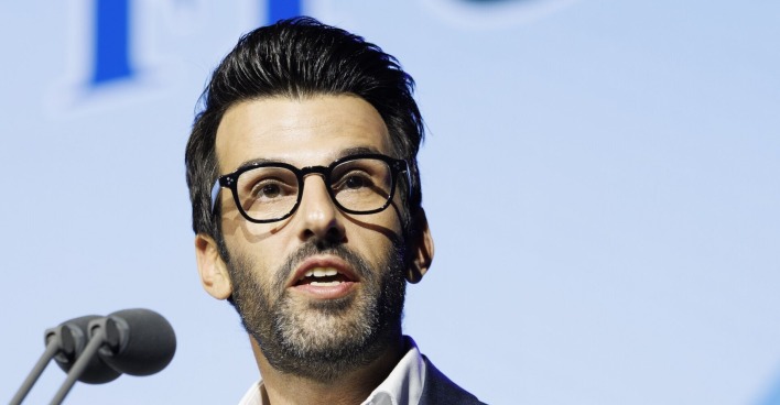 Udo Landbauer spricht vor Mikrofon bei Pressekonferenz, trägt Brille und Anzug, Hintergrund mit unscharfem FPÖ-Logo.