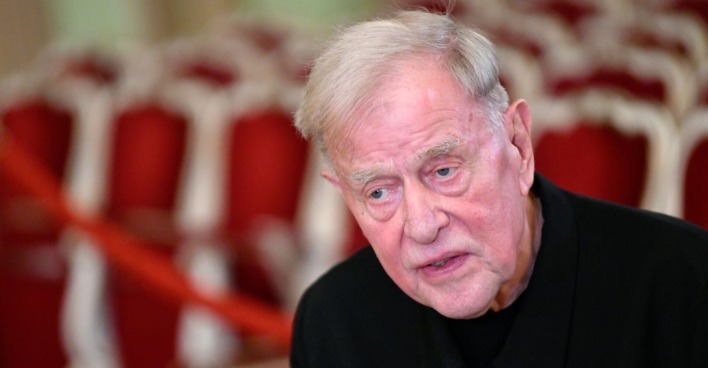 Der gealterte Claus Peymann mit ernstem Blick vor roten Theatersesseln – Großaufnahme des Regisseurs in einem Theatersaal.