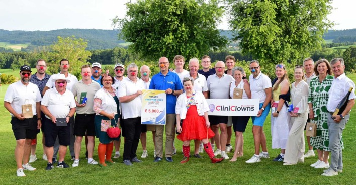 Gruppenfoto auf einem Golfplatz: Menschen mit roten Clownnasen halten einen 5.000 € Spendenscheck und ein CliniClowns-Schild.