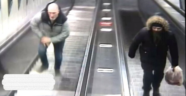 Fahndungsfoto: Zwei Männer auf Rolltreppen, links: ein Mann mit weißem Bart und dunkelgrüner Jacke, rechts: ein schwarz gekleideter, vermummter Mann mit Müllsack in der Hand und Kapuze über dem Kopf