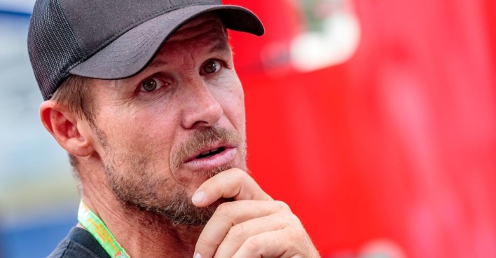Porträt von Felix Baumgartner mit schwarzer Kappe, nachdenklicher Gesichtsausdruck, im Vordergrund seine Hand am Kinn, unscharfer roter Hintergrund.