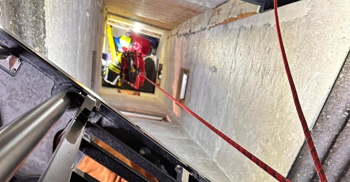 Blick in einen engen Schacht, in dem Feuerwehrleute mit einem roten Rettungskorb einen Jugendlichen nach oben ziehen. Ein rotes Sicherungsseil ist gespannt, eine Leiter ist im Vordergrund zu sehen.