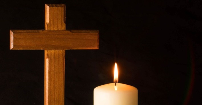 Eine leuchtende weiße Kerze mit einem Holzkreuz vor schwarzem Hintergrund als Zeichen für Trauer