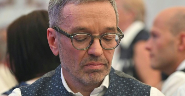 Herbert Kickl mit gesenktem Blick und ernstem Gesichtsausdruck, trägt Brille und Trachtenweste; im Hintergrund unscharf andere Personen.