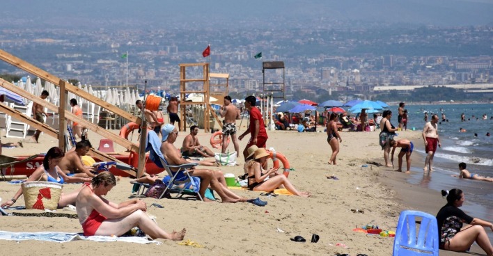 Menschen an einem überfüllten Sandstrand in Catania, Sizilien, bei extremer Sommerhitze; viele sitzen unter Sonnenschirmen oder gehen ins Meer zur Abkühlung.