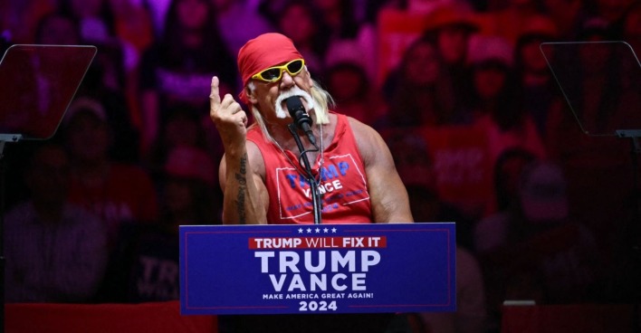 Hulk Hogan steht mit rotem Bandana und Sonnenbrille hinter einem Rednerpult mit der Aufschrift „Trump – Vance 2024“ und spricht bei einer Wahlkampfveranstaltung.