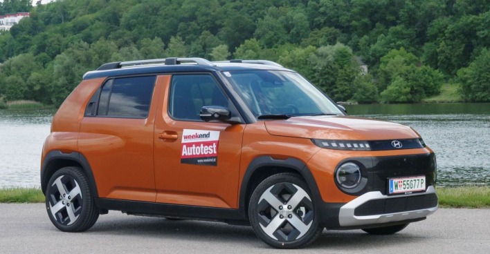 Ein kaffeebrauner Hyundai Inster parkt am Ufer eines Flusses. Am anderen Ufer ist ein dichter Laubwald zu sehen.