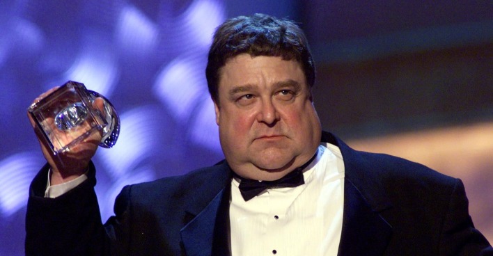 John Goodman früher stark übergewichtig im Smoking, mit ernstem Blick und Pokal in der Hand.