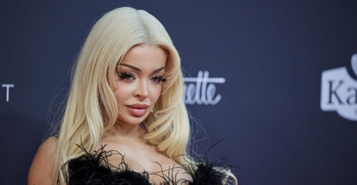 Katja Krasavice mit blondem Haar und tiefem Ausschnitt auf dem roten Teppich, schaut lasziv in die Kamera.