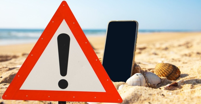 Warnschild und Handy im Sand am Strand: Symbolbild für Kostenfallen durch Handy-Nutzung im Urlaub.