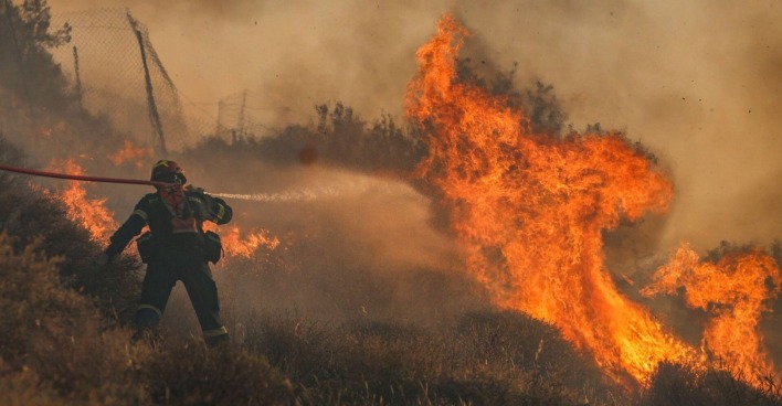 Ein Feuerwehrmann kämpft in Griechenland mit einem Wasserschlauch gegen meterhohe Flammen – dichter Rauch und Hitze erschweren den Einsatz massiv.