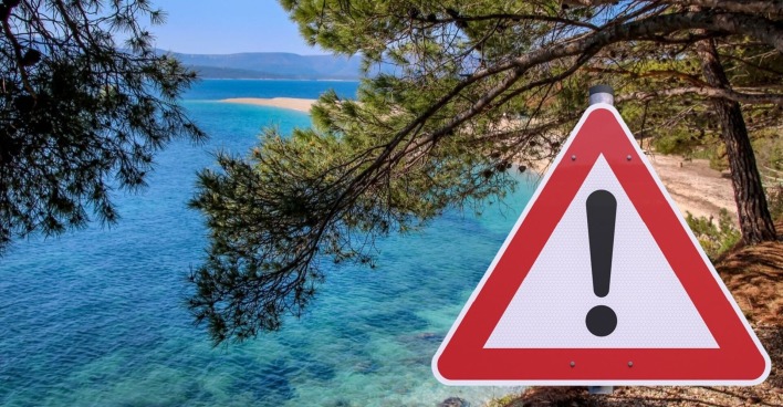 Warnschild mit Ausrufezeichen vor türkisblauem Meer und Pinien in Kroatien – Symbol für aktuelle Reisewarnungen wegen Waldbränden und Verkehrschaos.