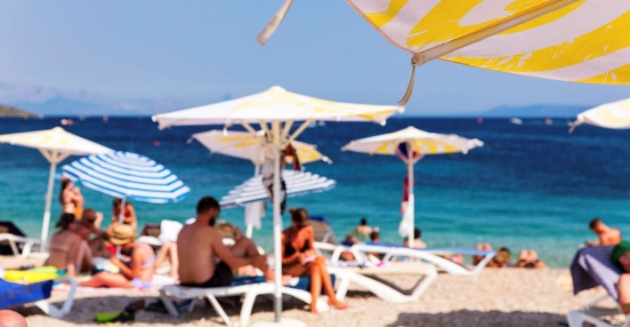 Aufnahme eines mediterranen Strandes mit unscharf zu erkennenden Badegästen, im Vordergrund ein Sonnenschirm