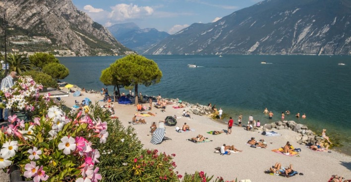 Menschen liegen auf Handtüchern und baden am Strand von Limone sul Garda, mit Blick auf den Gardasee und umliegende Berge, Blumen im Vordergrund