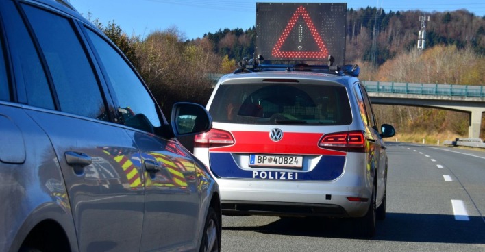 Polizeifahrzeug mit Warnanzeige und ein weiteres Auto stehen auf einer abgesperrten Landstraße in Österreich, im Hintergrund eine Brücke und Bäume.