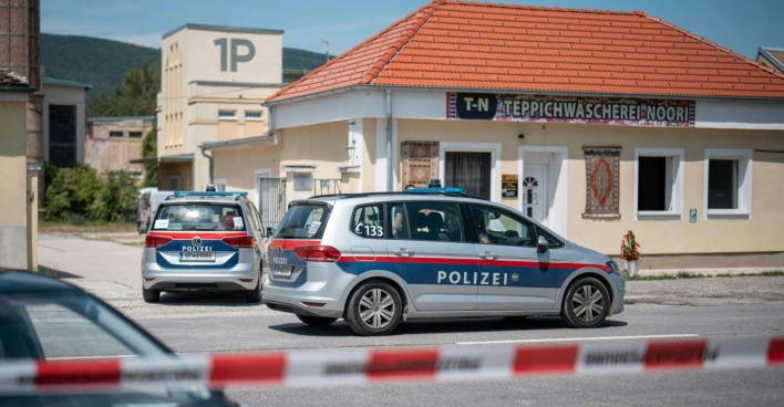 Zwei österreichische Polizeiautos stehen vor einem Gebäude in Traiskirchen mit der Aufschrift „Teppichwäscherei Noori“. Der Bereich ist mit rot-weißem Absperrband gesichert. Im Hintergrund sind Industriegebäude und grüne Hügel zu sehen.