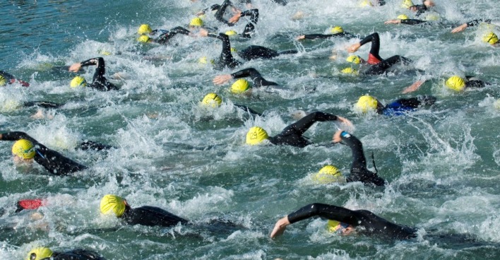 Zahlreiche Triathlet:innen mit gelben Badekappen und schwarzen Neoprenanzügen schwimmen eng nebeneinander im offenen Wasser eines Sees. Die Szene zeigt starke Wasserbewegung und Wettkampf-Stimmung.