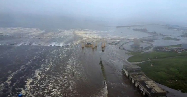 Luftaufnahme einer überfluteten Küstenstadt bei starkem Wellengang: Eine breite Tsunami-Welle überschwemmt Straßen, Häuser und Hafenanlagen unter grauem Himmel.