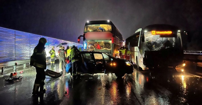 Unfallstelle bei Nacht auf der A10 Tauernautobahn: Zwei Busse und ein stark beschädigter Pkw stehen nach einem Crash auf regennasser Straße, Feuerwehrleute und Einsatzkräfte sichern die Unfallstelle.