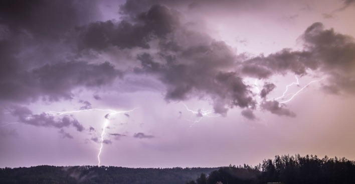 Gewitterfront mit dunklen Wolken und Blitzen über bewaldeter Landschaft