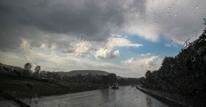 Blick durch eine regennasse Windschutzscheibe auf eine Landstraße, dunkle Gewitterwolken ziehen auf, dazwischen zeigen sich noch blaue Himmelsflecken.