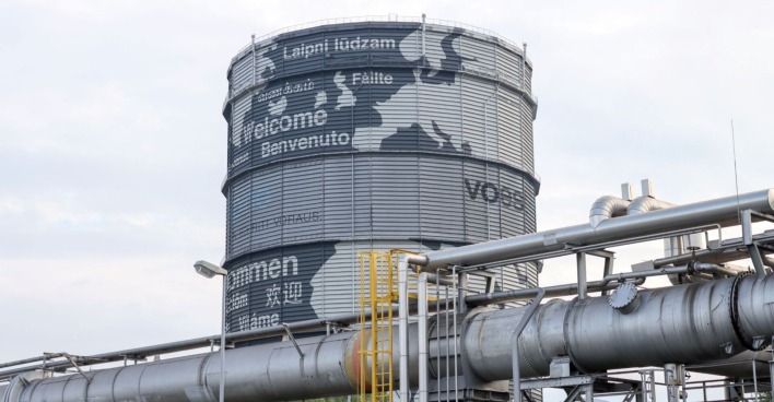 Großer, runder Gasspeicher bei der voestalpine in Linz mit internationalen Willkommensgrüßen, umgeben von industriellen Rohrleitungen.