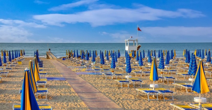 Leerer Strandabschnitt in Jesolo mit blauen und gelben Sonnenschirmen, Blick auf das Meer und einen Rettungsturm unter roter Flagge