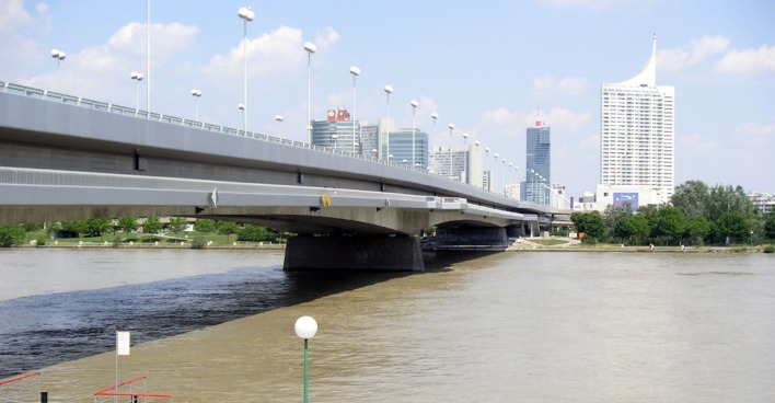Blick auf die Reichsbrücke in Wien über der Donau, im Hintergrund moderne Hochhäuser der Donau City, vorne Wasser mit sichtbarer Strömungsgrenze.