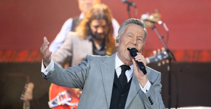 Roland Kaiser singt live auf der Bühne, im grauen Anzug mit Mikrofon in der Hand, Musiker im Hintergrund.