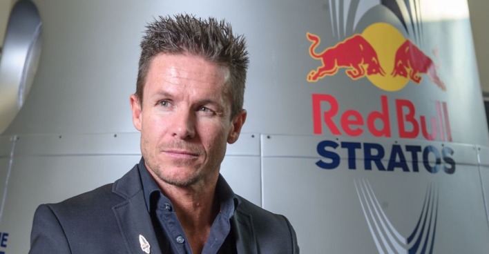 Felix Baumgartner steht in einem dunklen Anzug vor der Red Bull Stratos-Kapsel mit dem Red Bull Logo im Hintergrund.