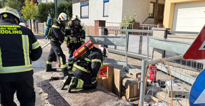 Mehrere Feuerwehrleute in Schutzkleidung und mit Atemschutz arbeiten an einer abgesicherten Baugrube auf einer Straße in Mödling nach einem Gasaustritt.