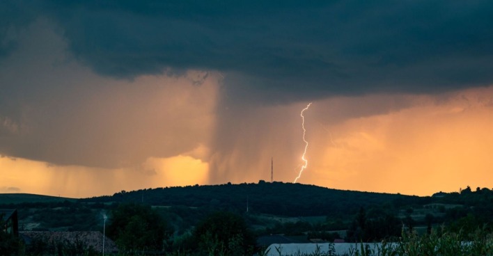 Dunkle Gewitterwolken über einer Hügellandschaft, ein Blitz schlägt neben einem Funkturm ein, im Vordergrund sind Häuser und Felder zu sehen.