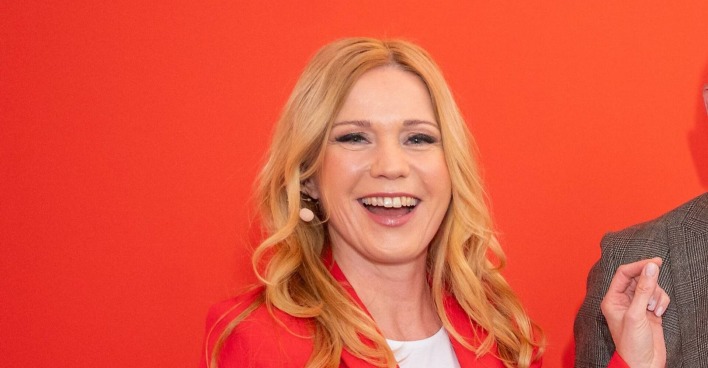Moderatorin Johanna Setzer, mit langen blonden Haaren lächelt in rotem Blazer vor rotem Hintergrund.