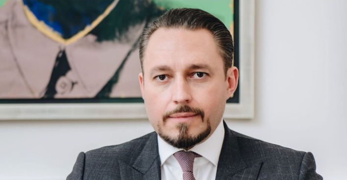 Ein Mann in Anzug und Krawatte blickt direkt in die Kamera. Er hat streng zurückgekämmtes Haar, einen gepflegten Bart und ernsten Gesichtsausdruck. Im Hintergrund ist ein modernes Gemälde zu sehen.