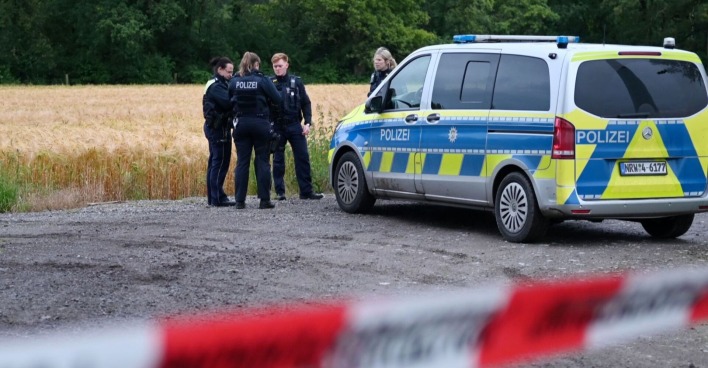 Mehrere Polizistinnen und Polizisten stehen neben einem Einsatzfahrzeug vor einem abgeernteten Feld, eine rot-weiße Absperrung ist im Vordergrund zu sehen.