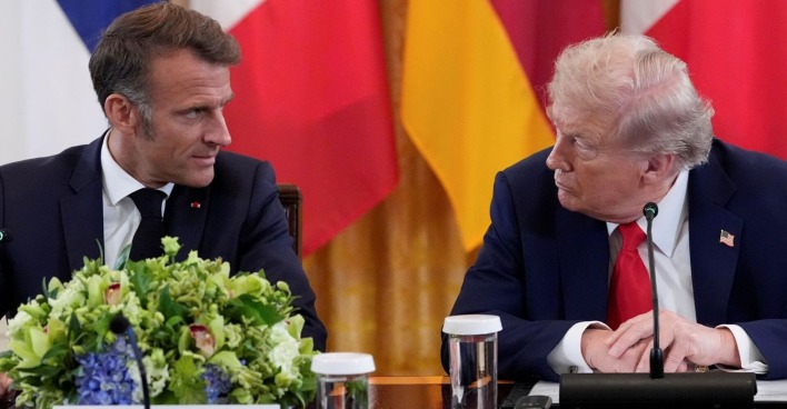 Frankreichs Präsident Emmanuel Macron und US-Präsident Donald Trump sitzen nebeneinander an einem Konferenztisch im Weißen Haus. Beide blicken sich ernst an, vor ihnen stehen Mikrofone, Gläser und ein großer Blumenschmuck. Im Hintergrund sind Flaggen verschiedener Länder zu sehen.