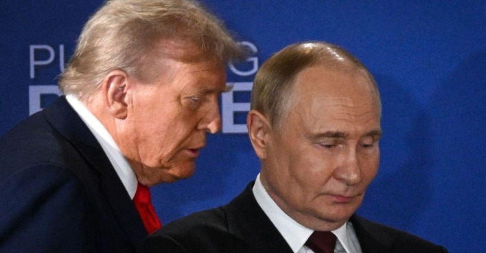 Donald Trump spricht dicht neben Wladimir Putin, der mit ernster Miene nach unten blickt. Beide tragen Anzug, im Hintergrund eine blaue Wand mit dem Schriftzug "Pursuing Peace".