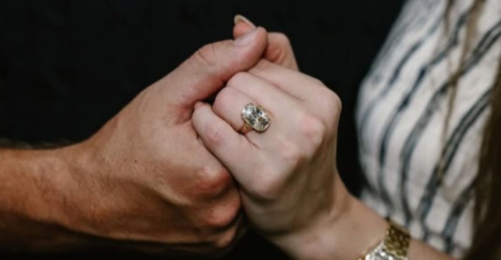 Nahaufnahme zweier sich haltender Hände, im Fokus ein großer rechteckiger Diamantring mit Gelbgoldfassung am Finger von Taylor Swift, die ein gestreiftes Kleid trägt.