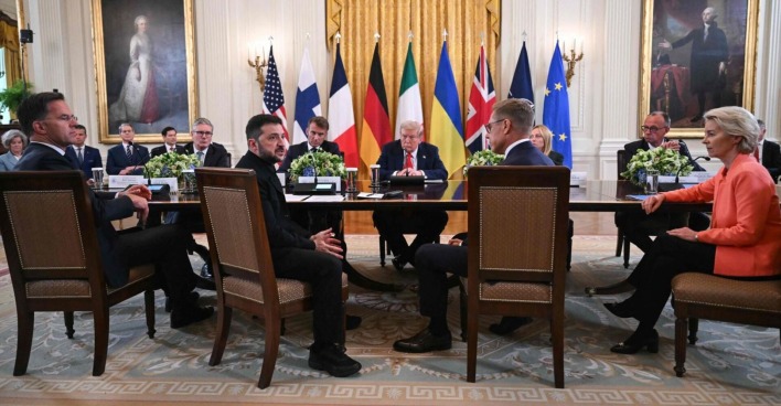 Ukraines Präsident Selenskyj, US-Präsident Trump und führende Politiker aus der EU und der NATO sitzen im Weißen Haus an einem großen Konferenztisch. Im Hintergrund stehen Landesflaggen, links und rechts hängen Porträts früherer Persönlichkeiten. Die Anwesenden blicken ernst, die Atmosphäre wirkt konzentriert.
