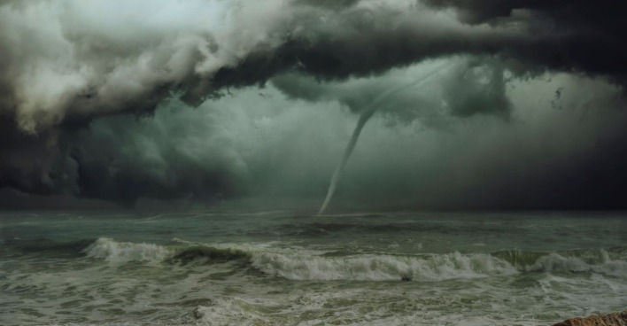 Dunkle Gewitterwolken über dem Meer mit einem gut sichtbaren Tornado, der sich aus der Wolkendecke bis zur Wasseroberfläche erstreckt. Im Vordergrund tobt die Brandung.