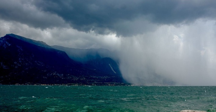 Dunkle Gewitterwolken und heftiger Starkregen über dem Gardasee. Der linke Bildteil zeigt dunkle Berge unter einer bedrohlichen Wolkendecke, rechts fällt dichter Regen über die Küste. Im Vordergrund ist aufgewühltes, türkisgrünes Wasser zu sehen.